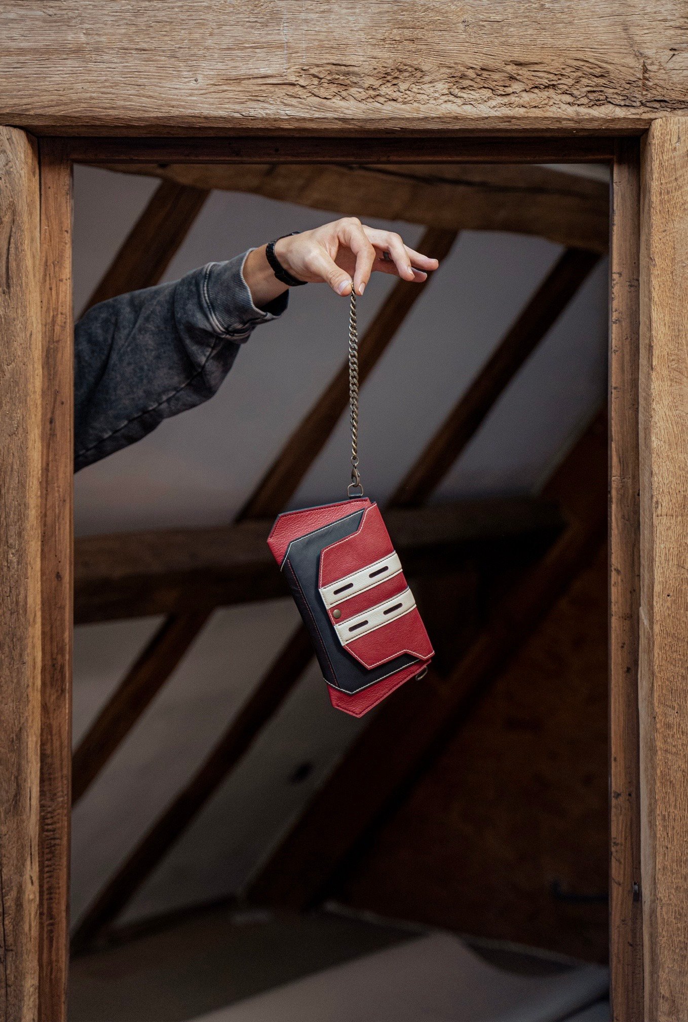Petite sacoche en cuir tricolore — rouge, noir et écru — suspendue à sa chaîne et tenue à la main, dans l'atelier aux poutres de bois